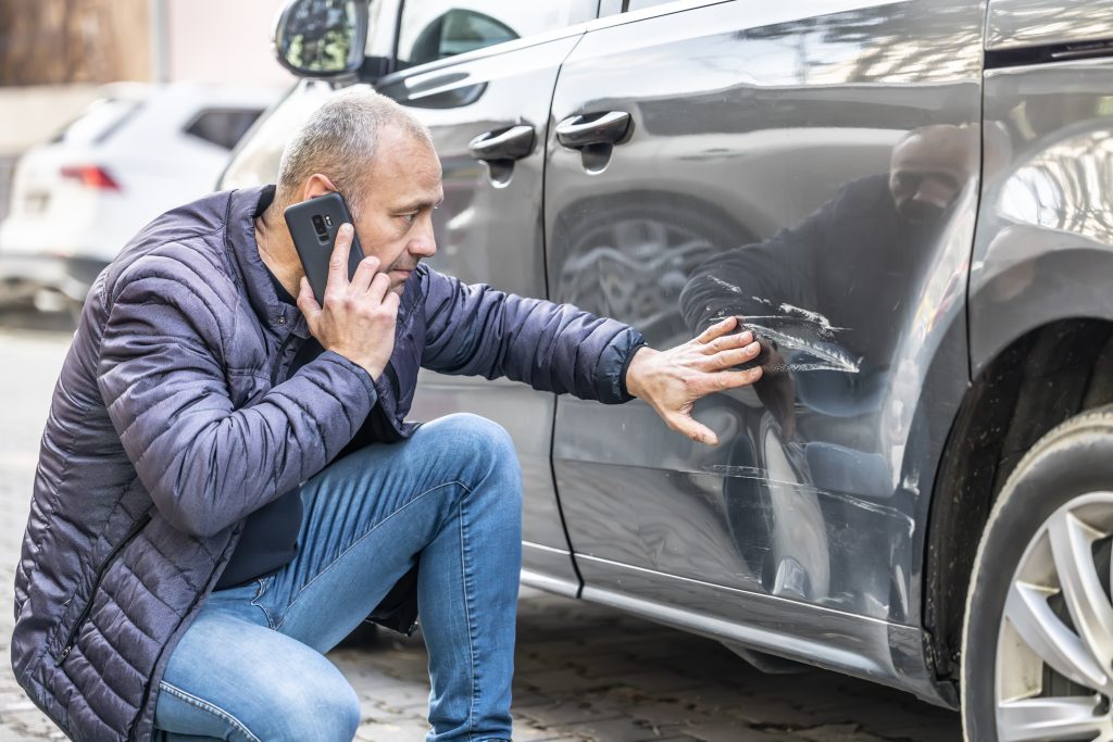 person on a cell phone looking at the left side of a car with body damage to the paint